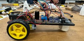 Selbstgebauter Roboter steht auf Werkbank. Im Hintergrund arbeiten Schüler*innen am Werkstück 