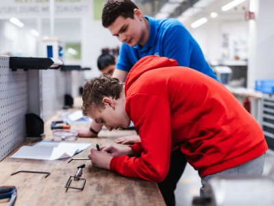 Ein Schüler und ein Azubis messen zusammen am Werktisch ein Bauelement aus