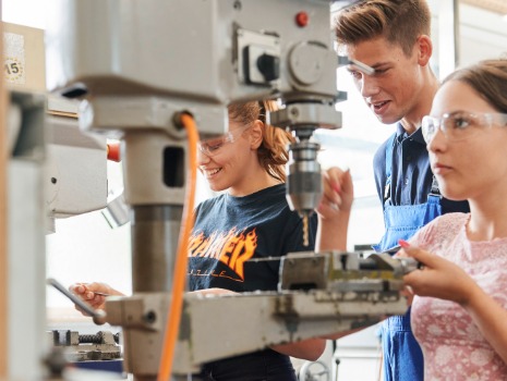 Azubi steht mit zwei Teilnehmenden vor einer Standbohrmaschine und gibt ihnen Anweisungen 