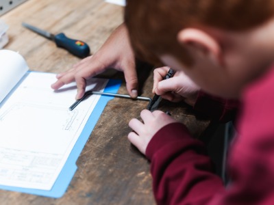 Schüler misst auf einer Werkbank mit Hilfe eines Azubis  ein Werkstück aus 